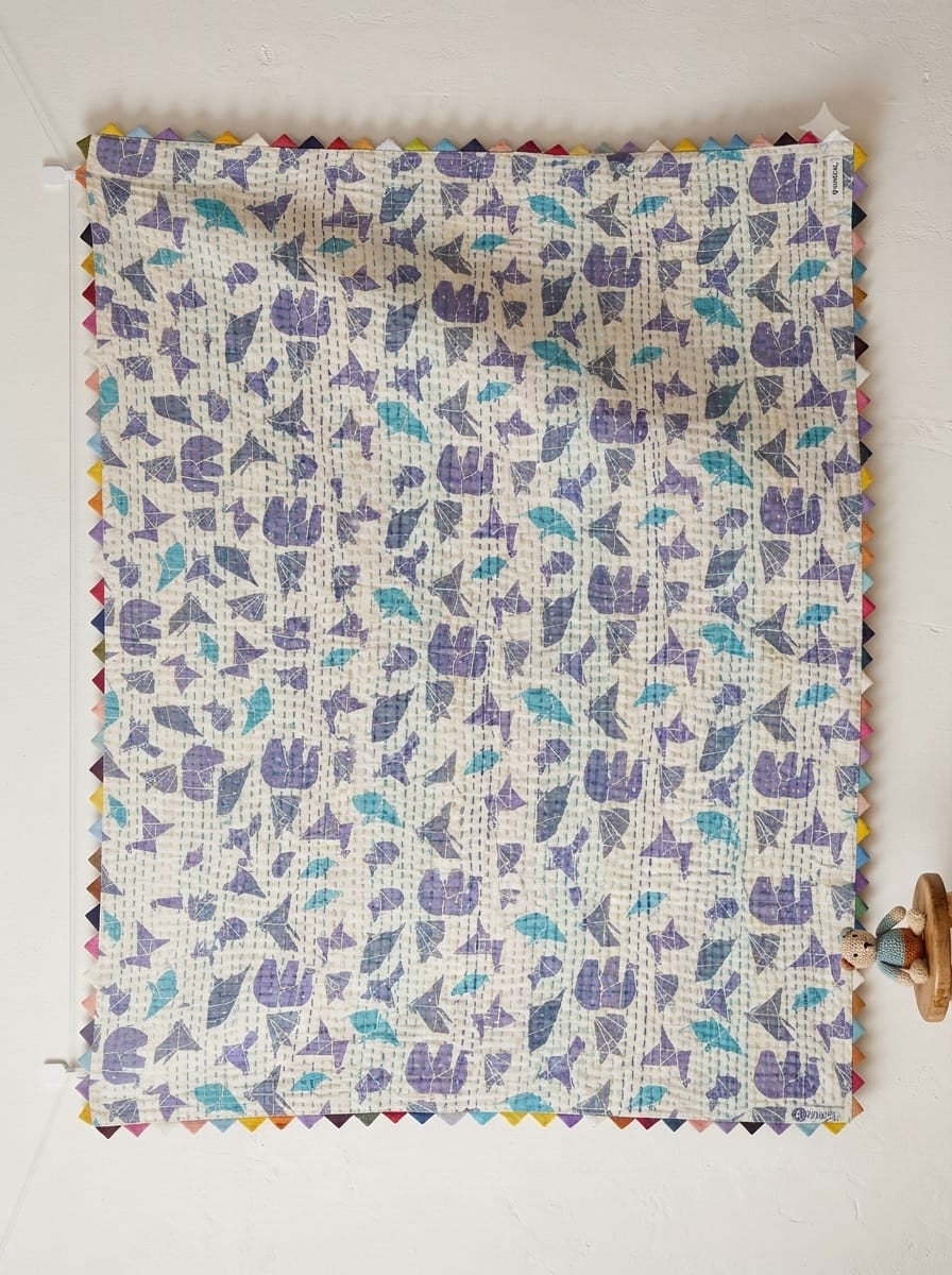 Hanging off-white cotton baby quilt featuring purple and teal origami animal motifs with hand-stitched Kantha quilting and colorful triangle edge binding.