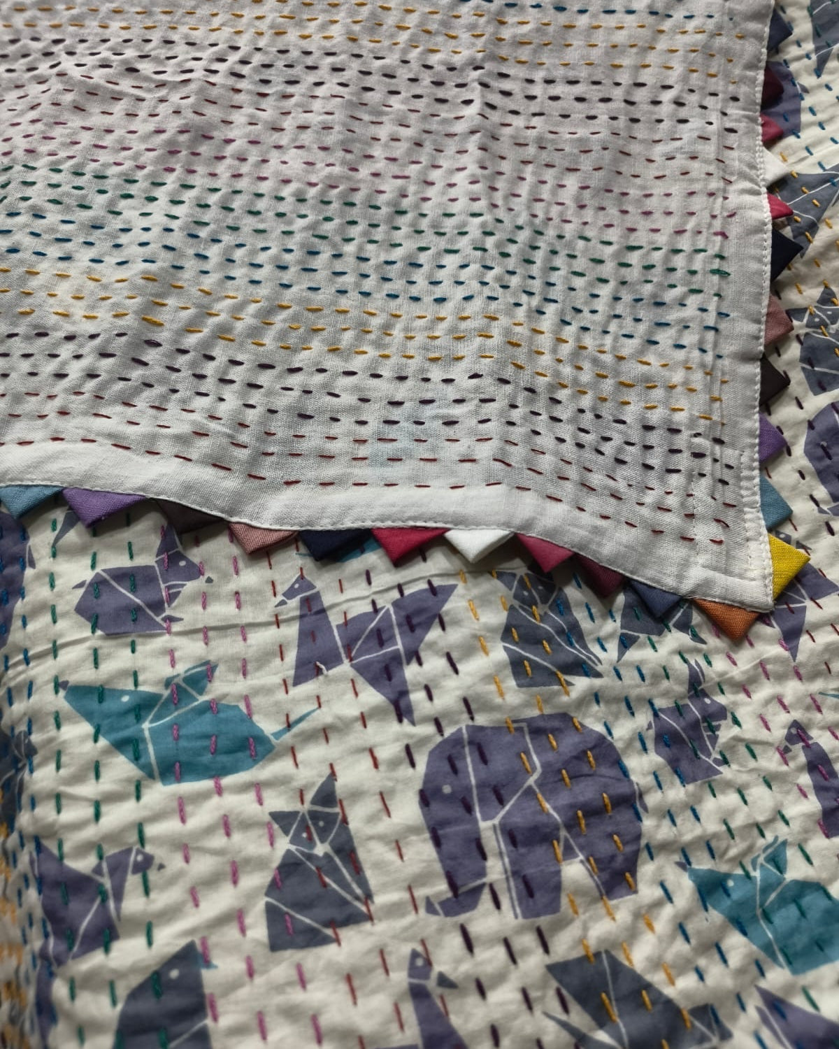 Hanging off-white cotton baby quilt featuring purple and teal origami animal motifs with hand-stitched Kantha quilting and colorful triangle edge binding.
