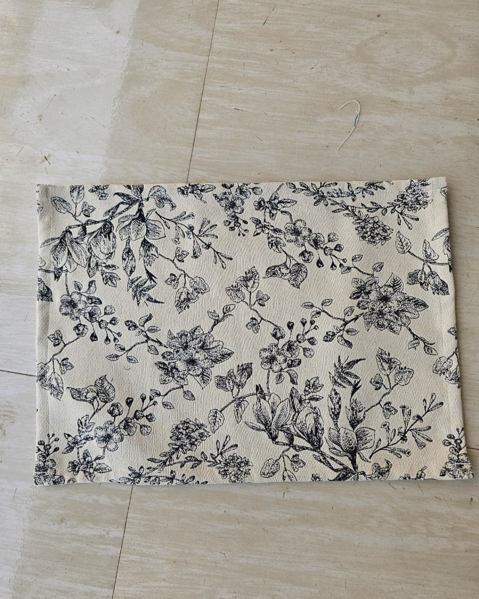 Rectangular off-white cotton placemat featuring a detailed black line-art floral botanical print laid flat.