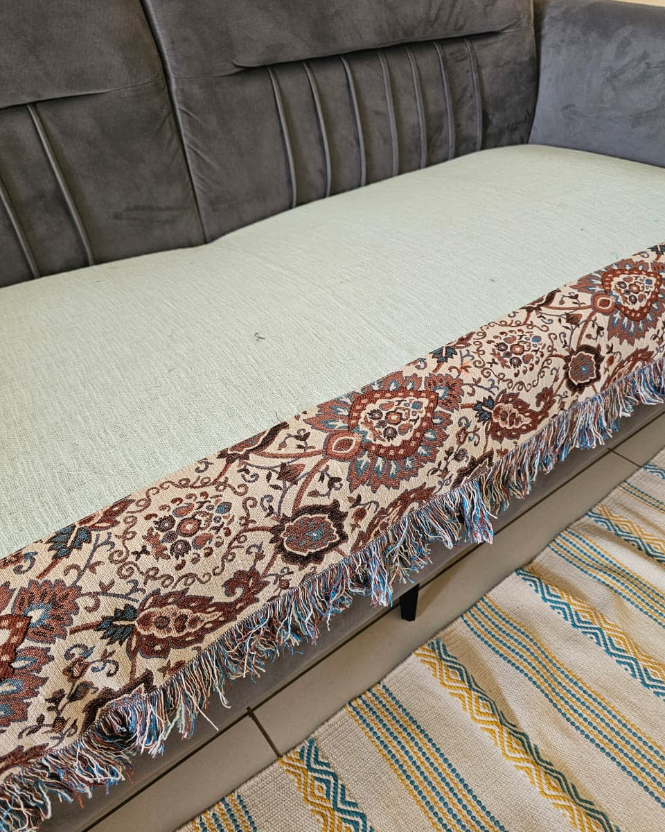 Light sage mint green jute cotton sofa seating cover featuring a vintage floral tapestry patchwork border with fringed tassels on a grey velvet sofa.
