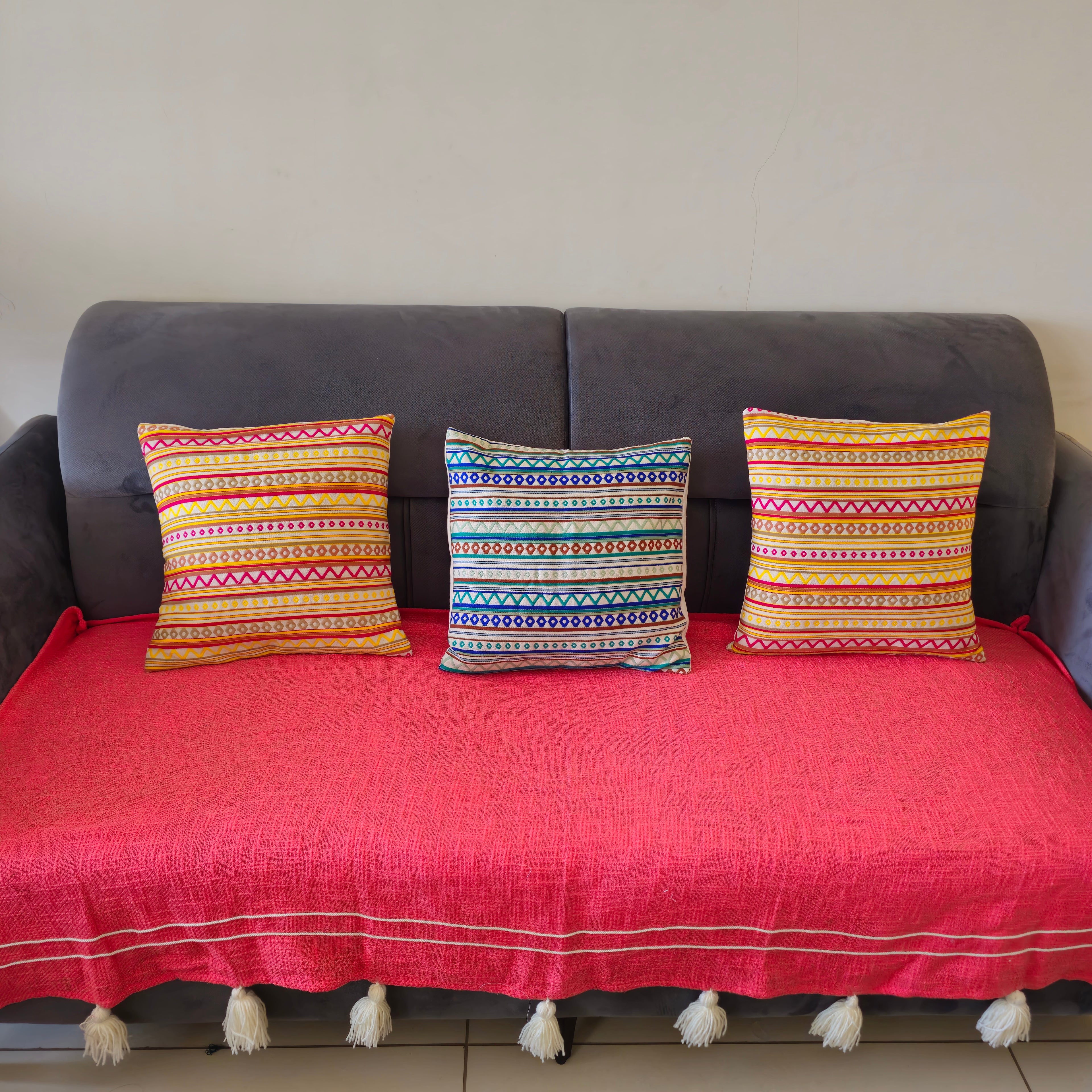 A pink slub cotton sofa cover with thread work and tassel detailing displayed on a sofa, accompanied by two colorful striped cushions.