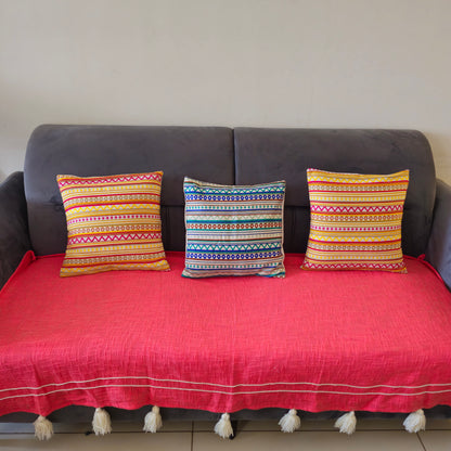 A pink slub cotton sofa cover with thread work and tassel detailing displayed on a sofa, accompanied by two colorful striped cushions.