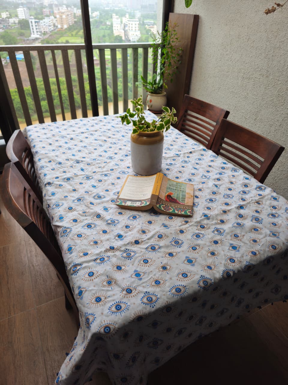 A rectangular dining table covered with a white tablecloth featuring a blue and green floral pattern, surrounded by dark wooden chairs. A small potted plant and an open book sit on the table, with a balcony railing and outdoor view in the background.