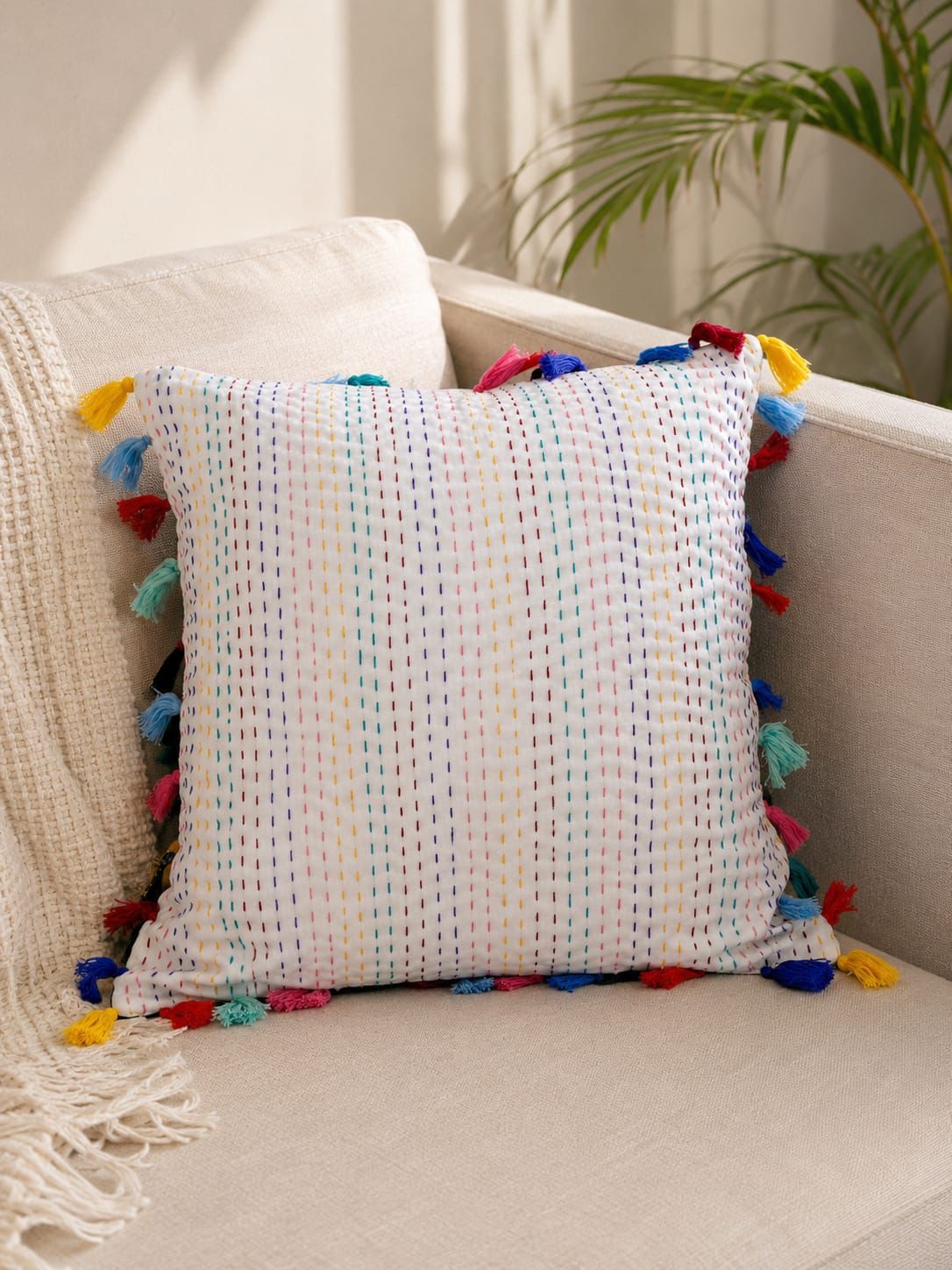 Square off-white cotton cushion cover with vertical multicolored Kantha hand stitching and vibrant rainbow tassels on a beige sofa.