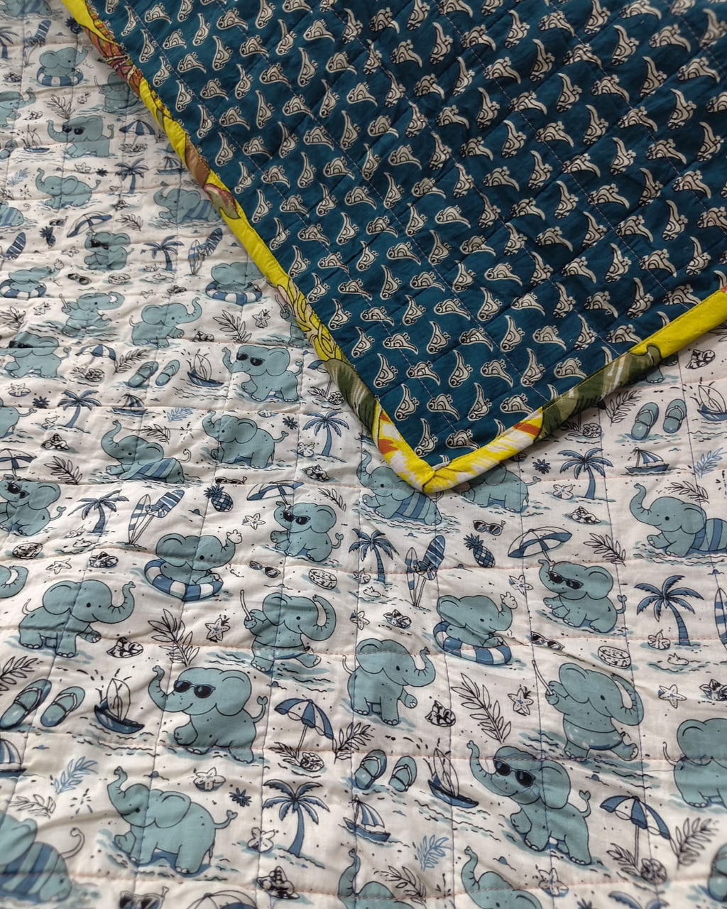 Reversible cotton baby quilt featuring a light blue beach elephant print with one corner folded over to reveal the deep teal geometric reverse side and sunny yellow binding.