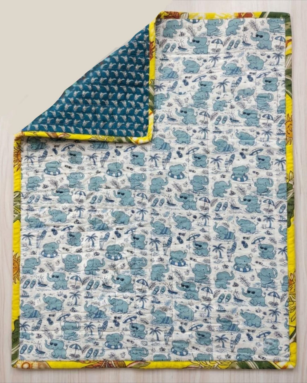 Reversible cotton baby quilt featuring a light blue beach elephant print with one corner folded over to reveal the deep teal geometric reverse side and sunny yellow binding.