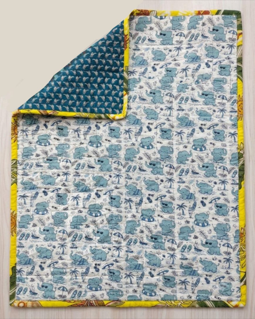 Reversible cotton baby quilt featuring a light blue beach elephant print with one corner folded over to reveal the deep teal geometric reverse side and sunny yellow binding.