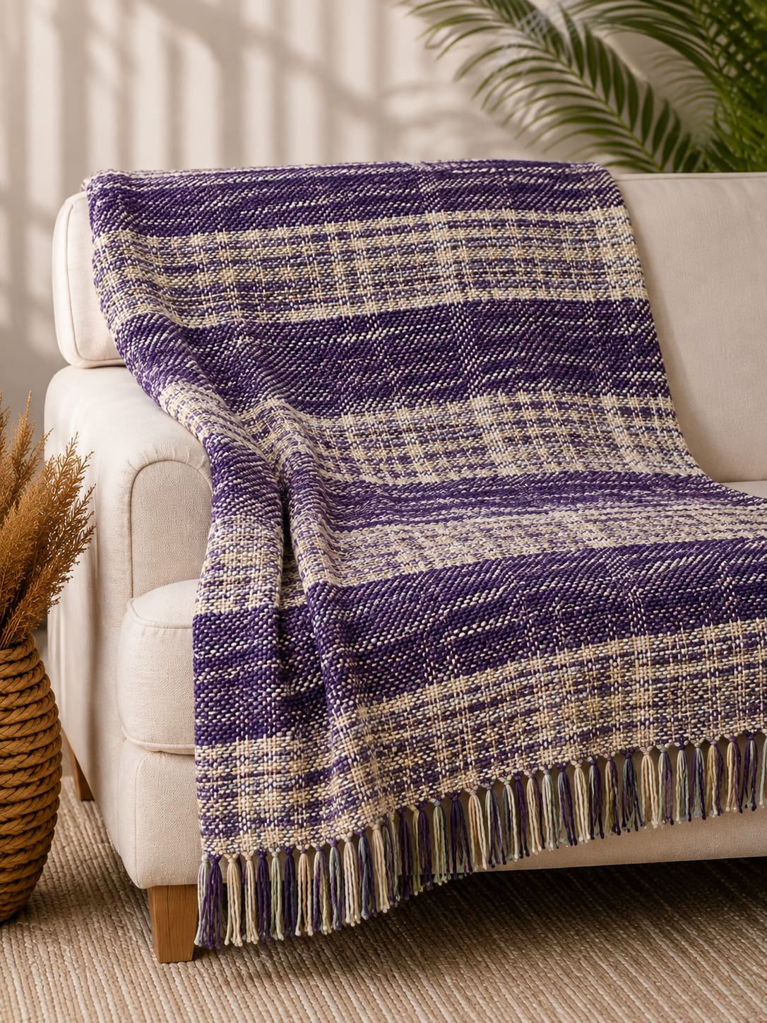 Royal purple and cream handwoven woolen sofa throw blanket with tasseled fringe draped over a cream-colored couch.