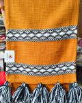 Rust orange jute-cotton handloom table runner featuring chunky off-white woolen hanging tassels and Vasaca brand labels.