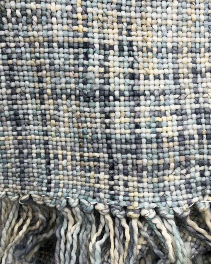 Close-up shot of a sage green and slate blue handwoven chunky woolen sofa throw featuring a textured plaid weave and thick twisted fringe tassels.