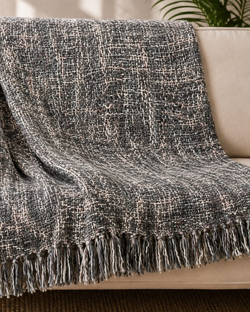Slate and charcoal grey handwoven chunky woolen sofa throw blanket featuring a textured tweed basketweave pattern and twisted fringe draped over a beige sofa.
