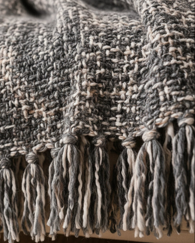 Slate and charcoal grey handwoven chunky woolen sofa throw blanket featuring a textured tweed basketweave pattern and twisted fringe draped over a beige sofa.