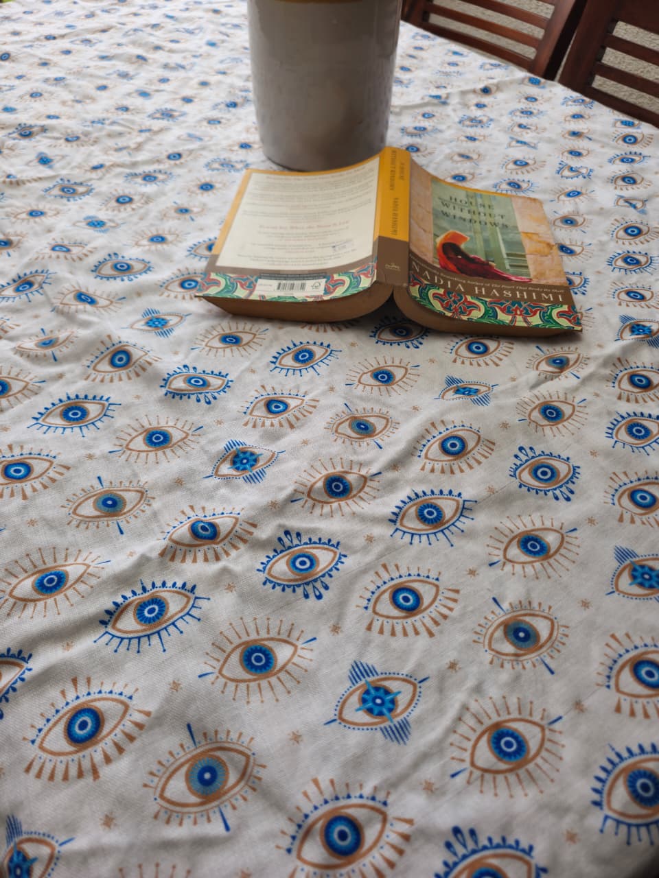 A close-up of a white tablecloth with a repeating hand-block printed pattern of blue and brown eyes, with an open book and a gray cup placed on it.