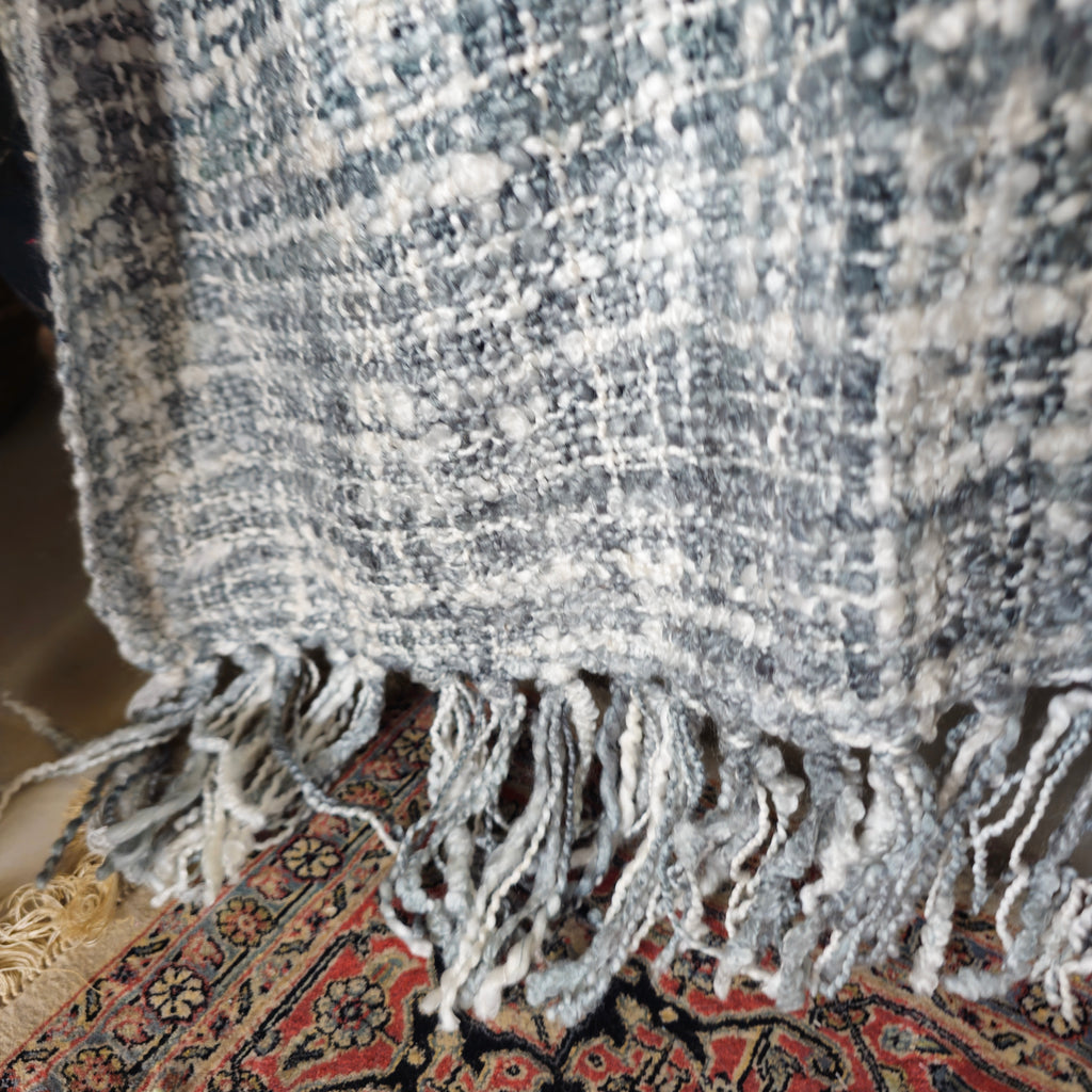 Close-up of a textured gray blanket with fringes on a patterned rug.