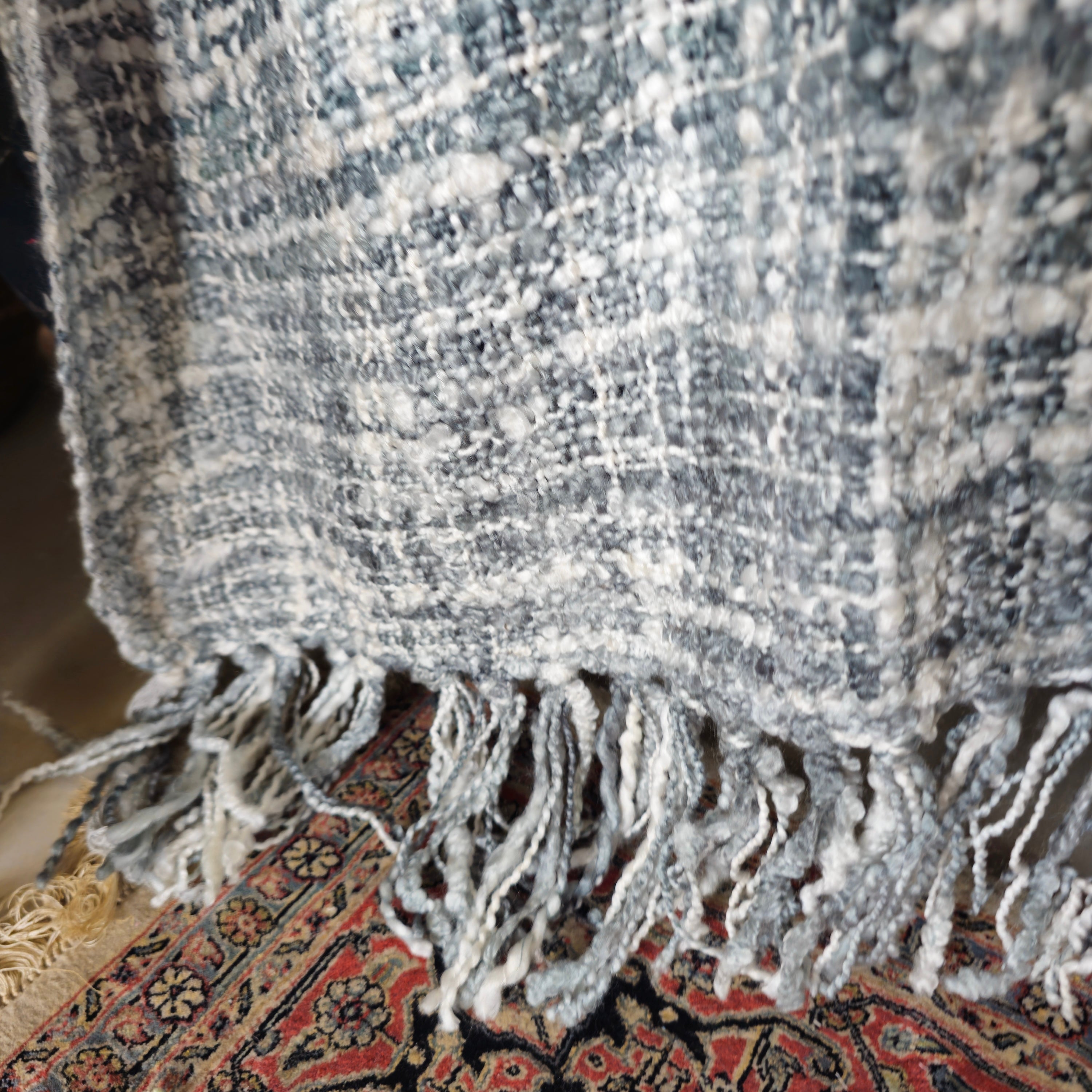 Close-up of a textured gray blanket with fringes on a patterned rug.
