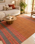 Terracotta rust cotton flatweave area rug with detailed charcoal grey and pink geometric border bands styled in a cozy warm living room.