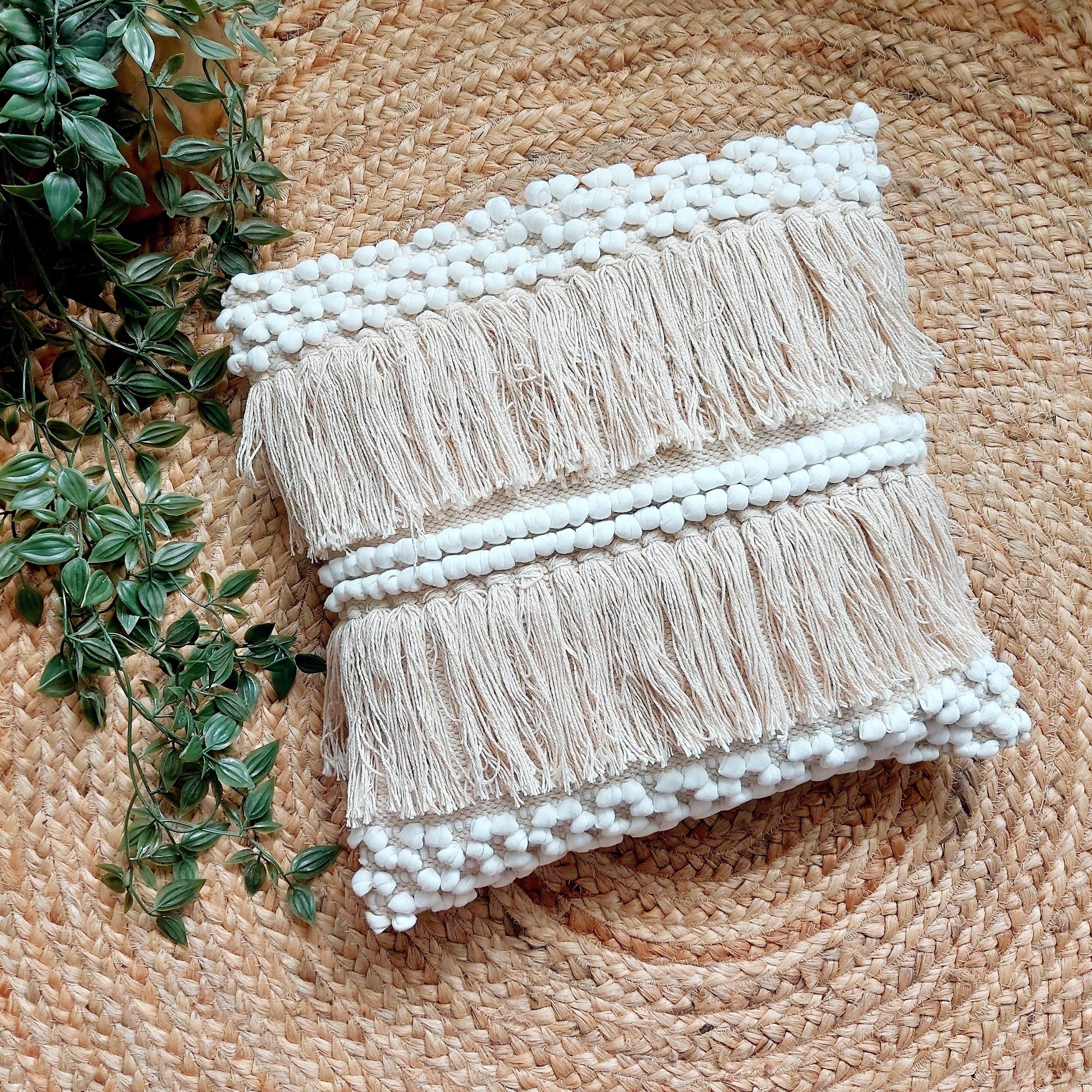 An off-white handwoven tufted boho cushion cover placed on a round rattan mat, with green foliage in the background.