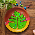Hand-painted terracotta wall plate featuring a vibrant green Monstera leaf over a pink and mustard yellow abstract background.