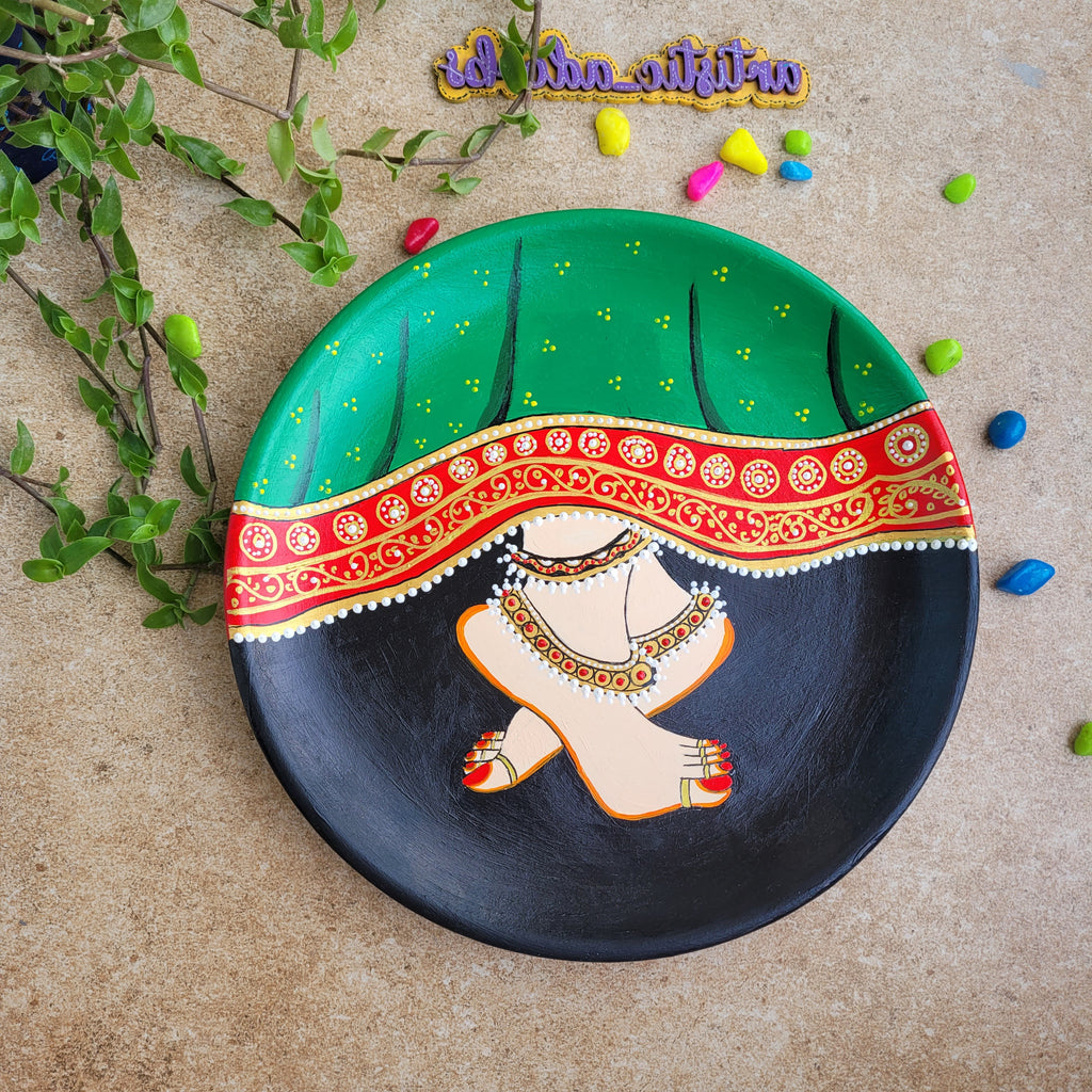 Hand-painted Pichwai terracotta wall plate detailing Radha's traditional anklet-clad feet beneath a patterned green lehenga skirt against a black backdrop.