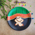 Hand-painted Pichwai terracotta wall plate detailing Radha's traditional anklet-clad feet beneath a patterned green lehenga skirt against a black backdrop.