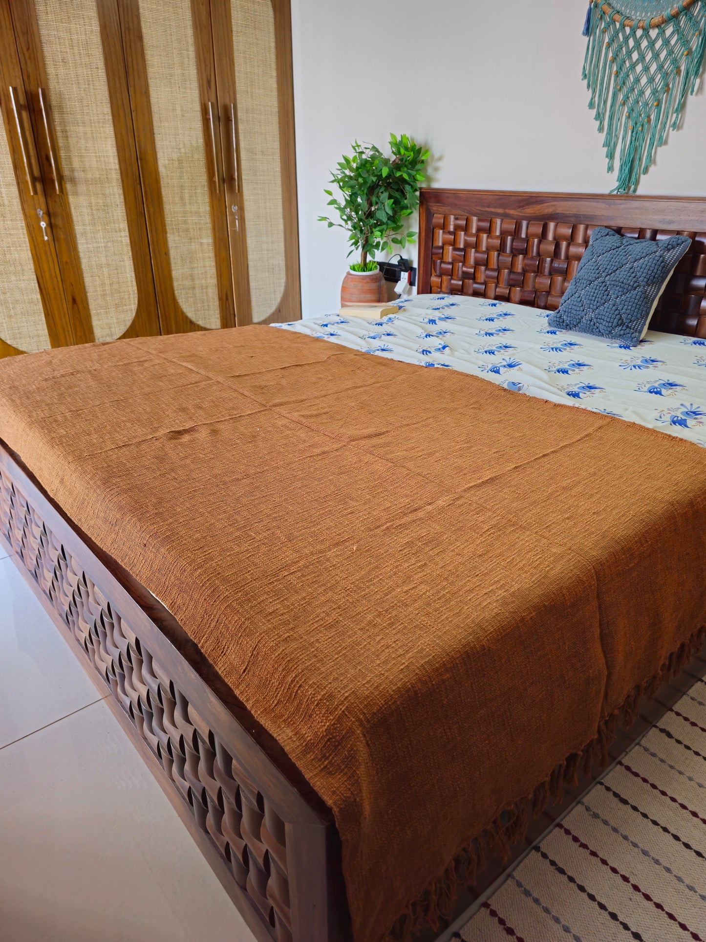 Bed with brown blanket and wooden headboard in a room with a plant and decorative wall hanging.