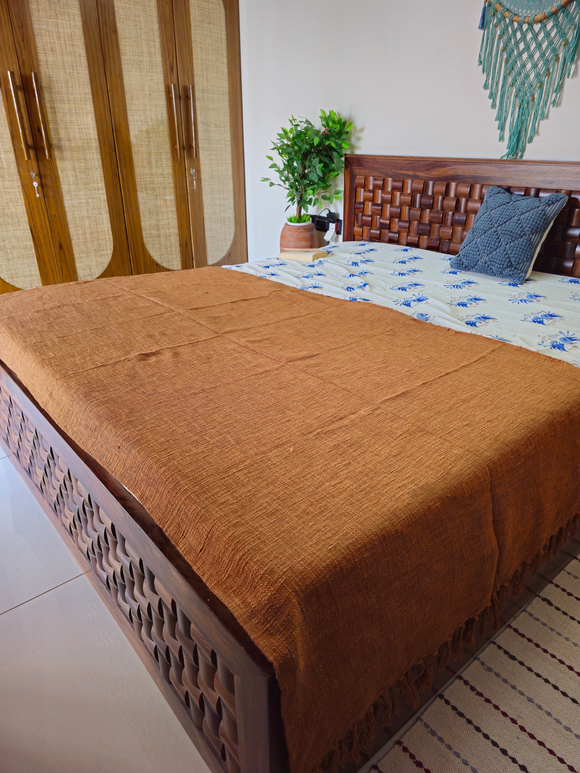 Bed with brown blanket and wooden headboard in a room with a plant and decorative wall hanging.