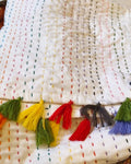 White cotton dining table runner featuring multicolored vertical running stitches and a colorful arrangement of yarn tassels along the edge, with Vasaca brand labels.