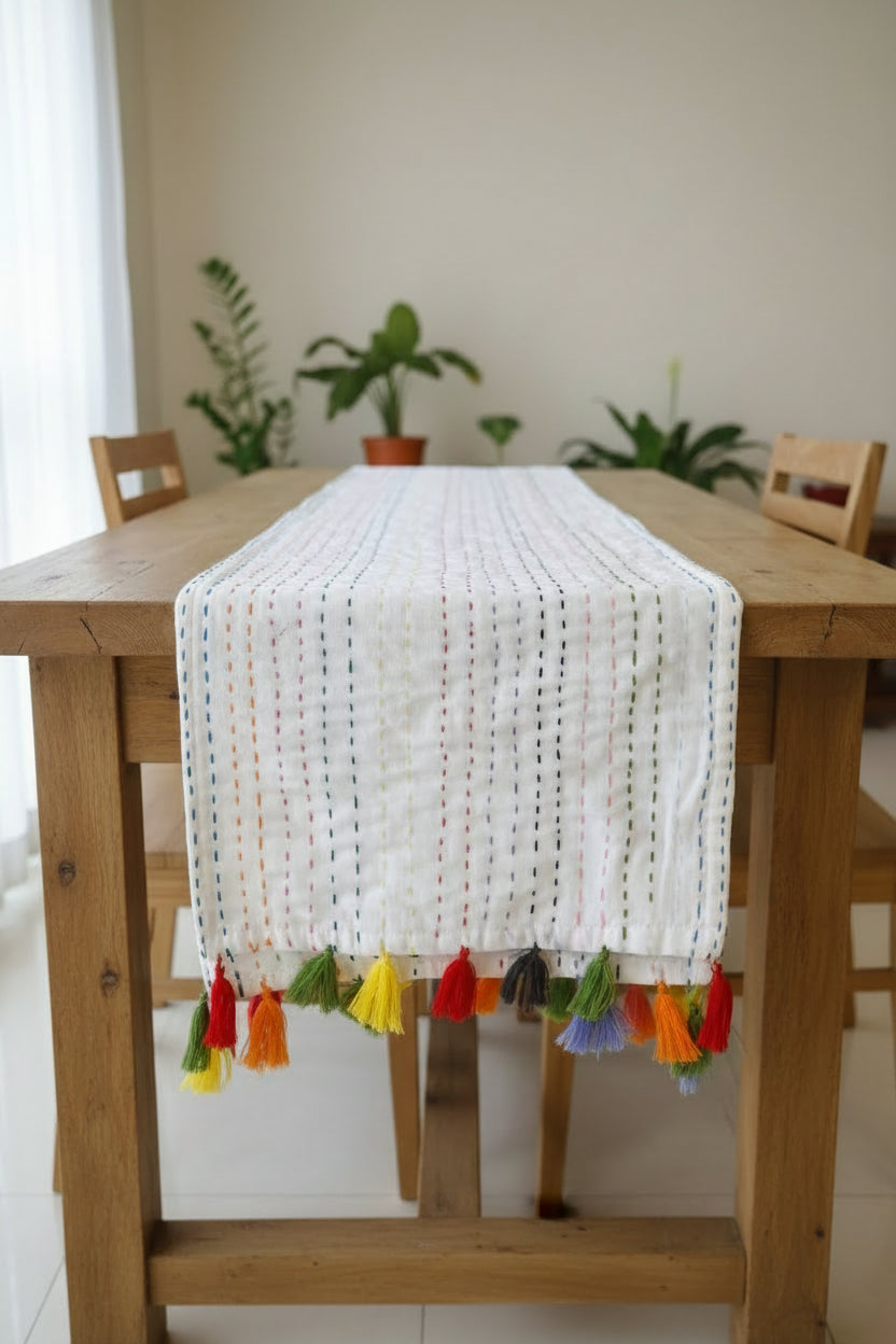 White cotton dining table runner featuring multicolored vertical running stitches and a colorful arrangement of yarn tassels along the edge, with Vasaca brand labels.