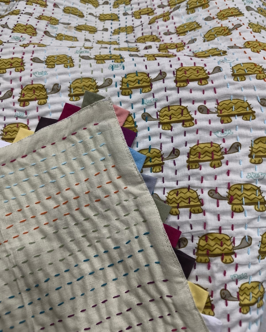 Close-up of a handmade reversible Kantha cotton baby quilt showing the solid neutral reverse side with multi-colored hand stitching folded over the yellow turtle print and colorful triangle edge binding.