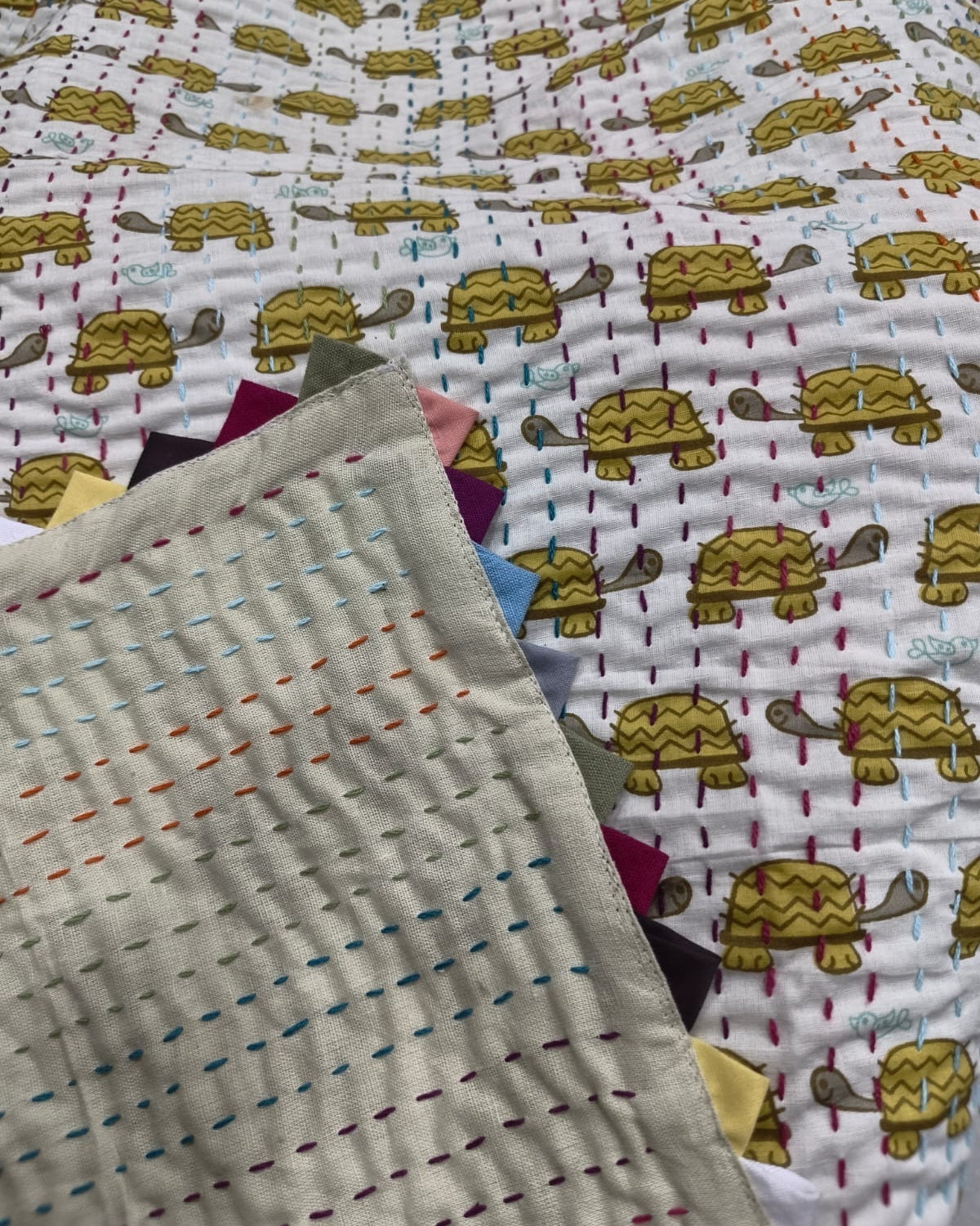 Close-up of a handmade reversible Kantha cotton baby quilt showing the solid neutral reverse side with multi-colored hand stitching folded over the yellow turtle print and colorful triangle edge binding.
