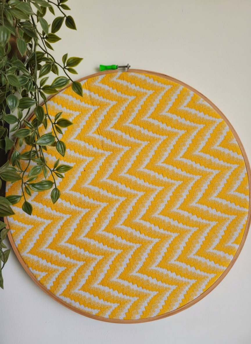 Round yellow and white zigzag patterned mat on a wooden frame against a white wall with a plant to the left.